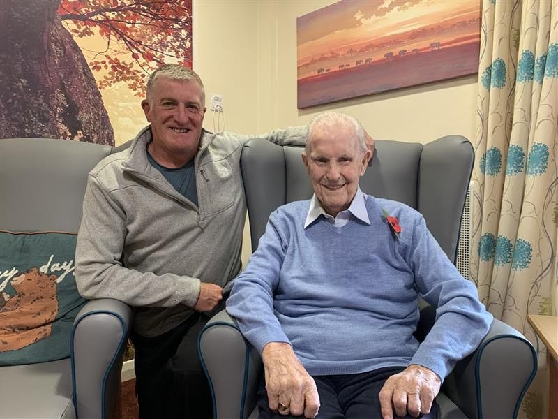 A young man standing next to Dennis Smith, 99-year old veteran, seated in an armchair